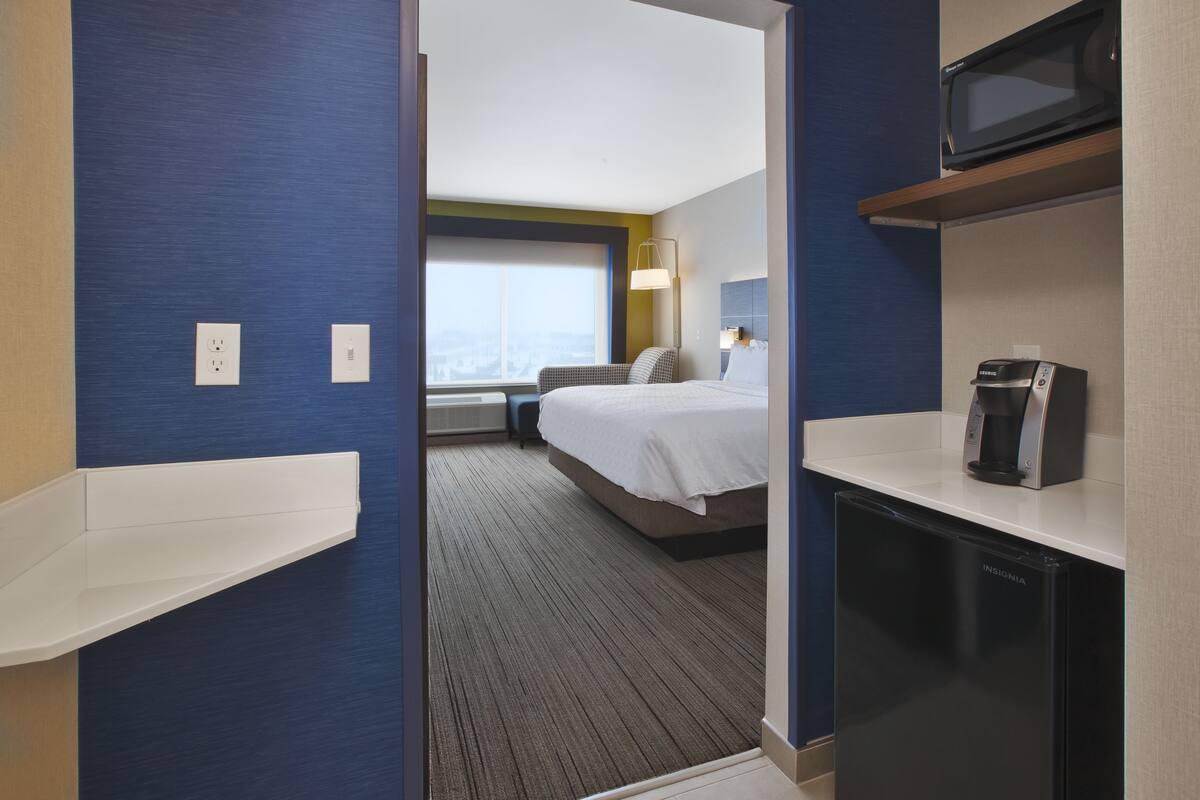 A hotel room with a bed, a microwave, a coffee maker, and a view of the city through the window.