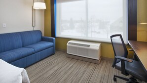 Une chambre d’hôtel avec un canapé bleu, un bureau en bois, une chaise de bureau noire, un lampadaire et une grande fenêtre donnant sur un paysage enneigé.