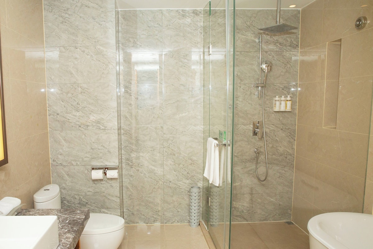 Un baño moderno con ducha envuelta en vidrio, paredes de mármol, un espejo grande y un lavamanos blanco.