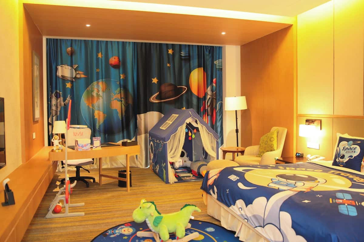 Habitación de hotel con cubrecama de temática espacial, escritorio con computadora, silla, área de juego tipo tienda de campaña y televisor.