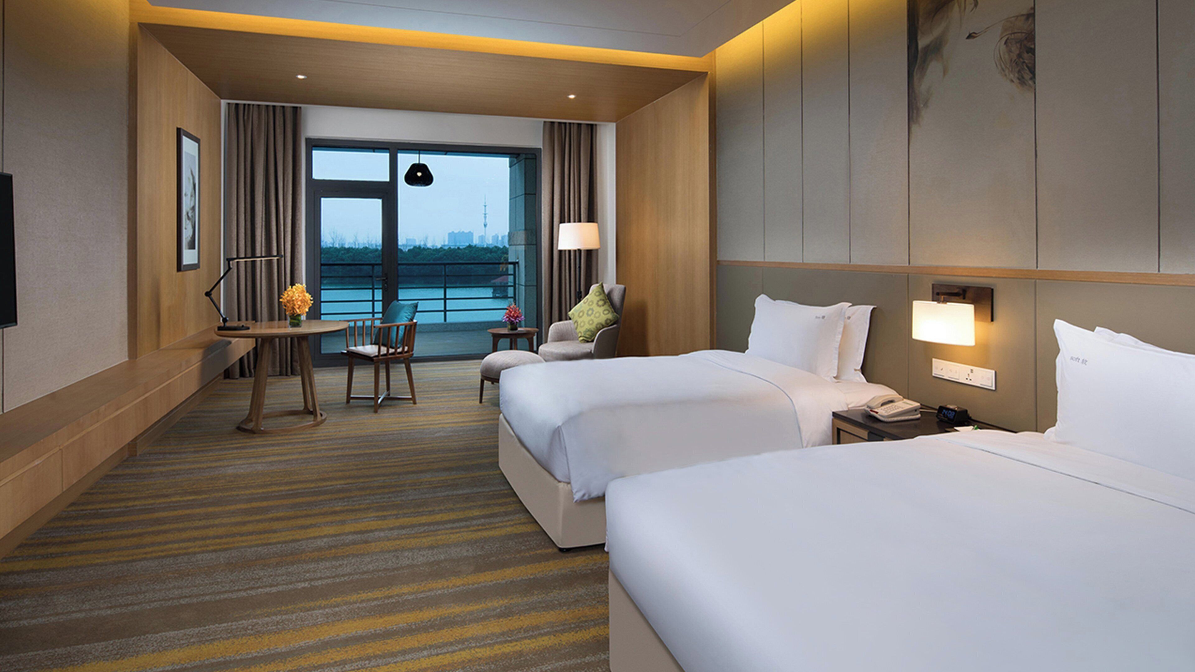 A hotel room with two beds, a desk, a chair, a lamp, and a view of the cityscape.