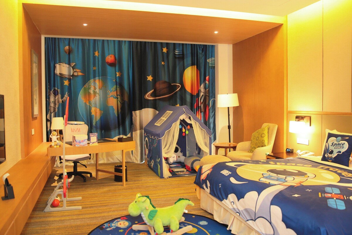 A hotel room with a space-themed bedspread, a desk with a computer, a chair, a small tent-like play area, and a television.