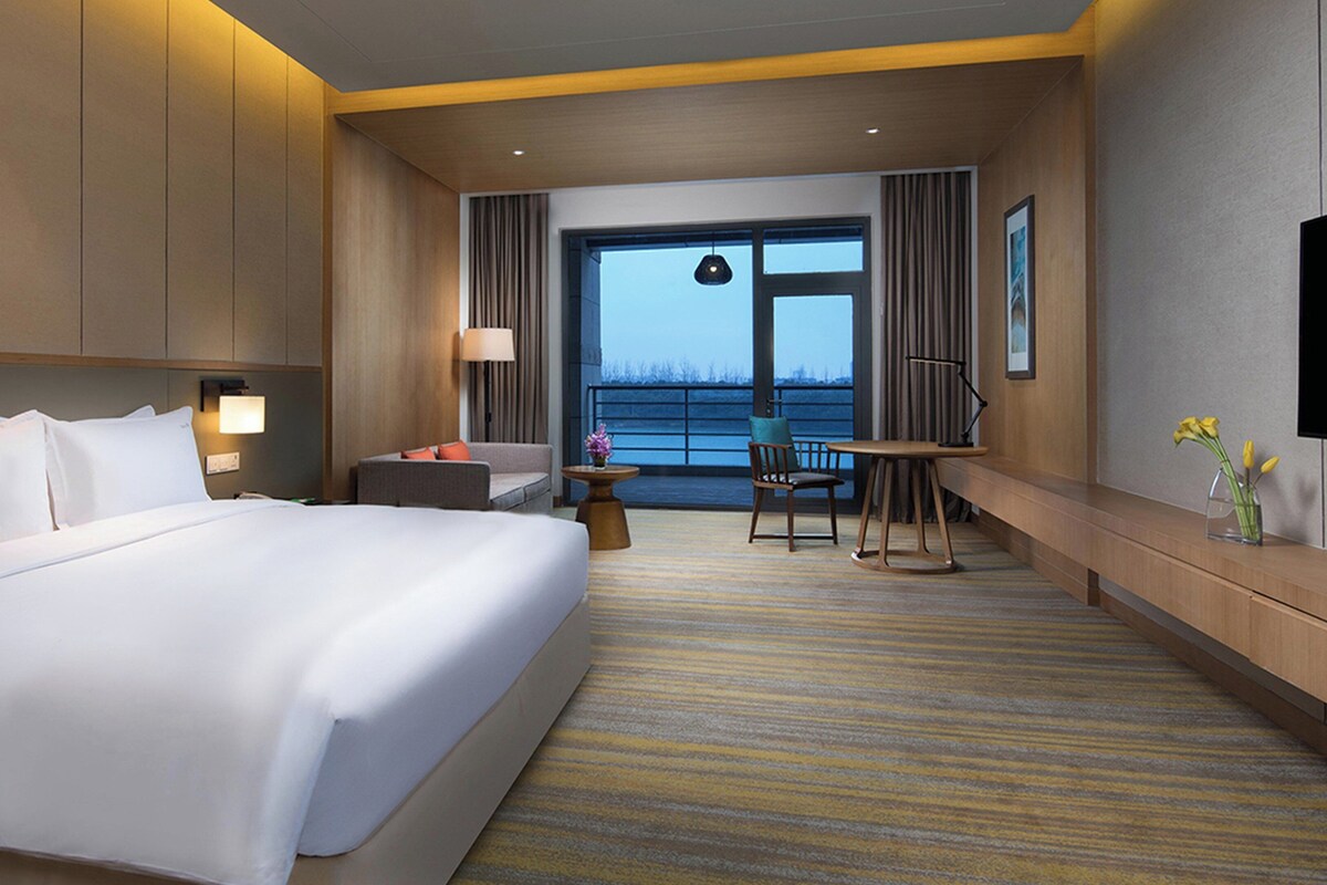 A hotel room with a large bed, a desk, a chair, and a view of the outdoors.