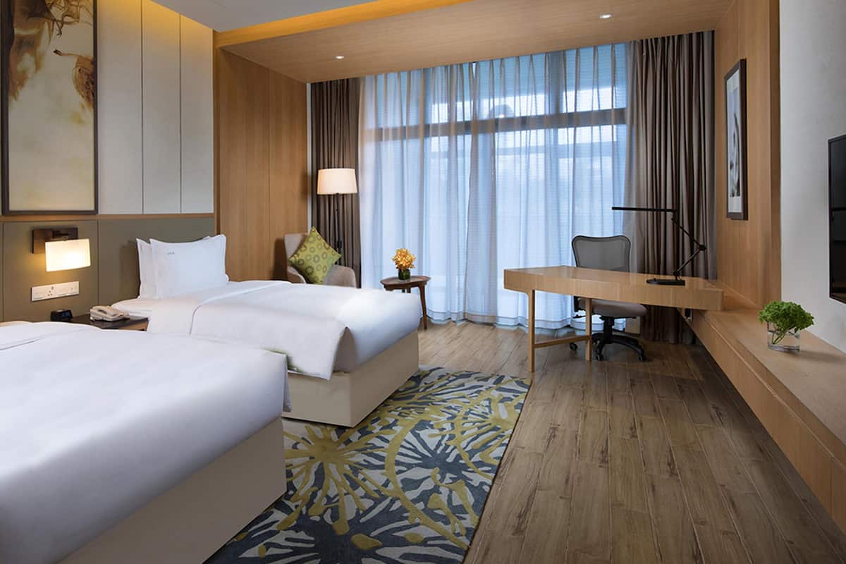 Habitación de hotel con dos camas, un escritorio, una silla, un televisor y un ventanal grande con cortinas.
