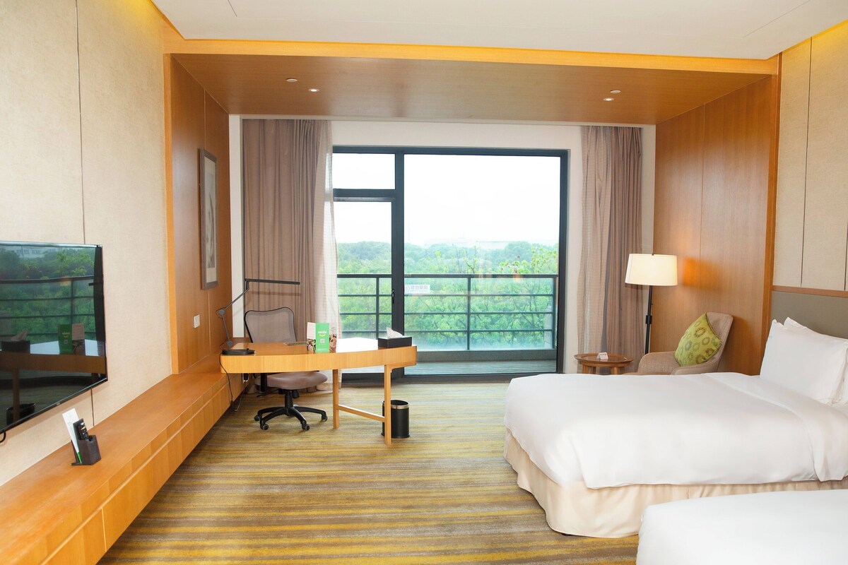 A hotel room with a bed, a desk with a chair, a TV mounted on the wall, and a window with curtains.