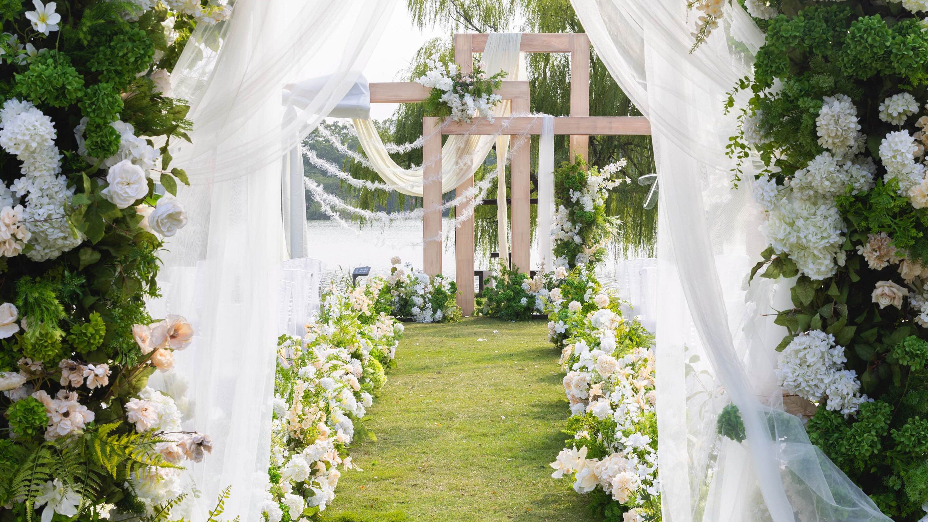 A wedding arch with white flowers and greenery, a grassy aisle, and a wooden structure in the background.