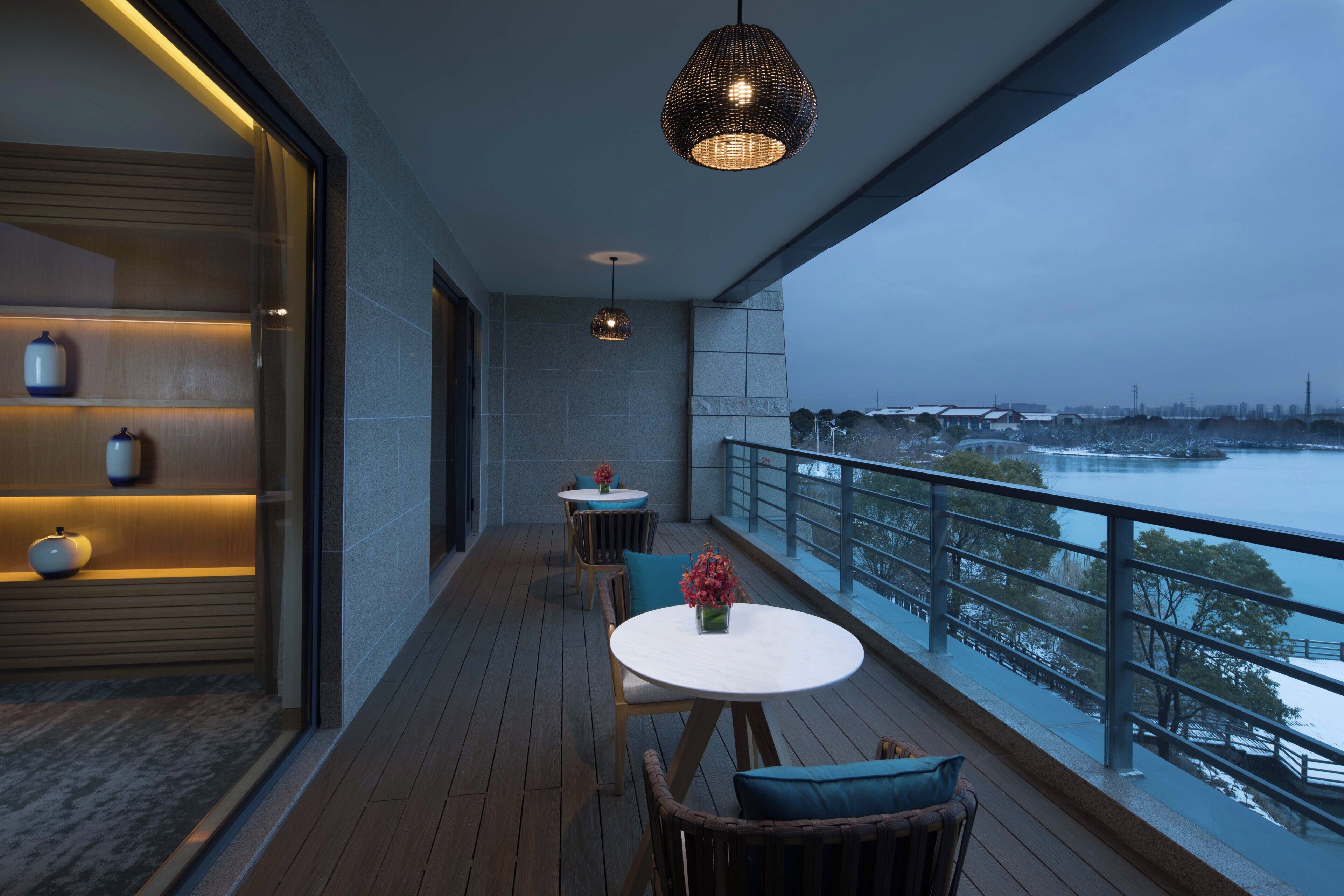 A modern balcony with a view of a river and cityscape, featuring a wooden floor, two tables with chairs, and decorative vases.