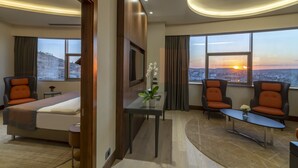 Een hotelkamer met een bed, twee fauteuils, een tafeltje en uitzicht op de ondergaande zon door grote ramen.