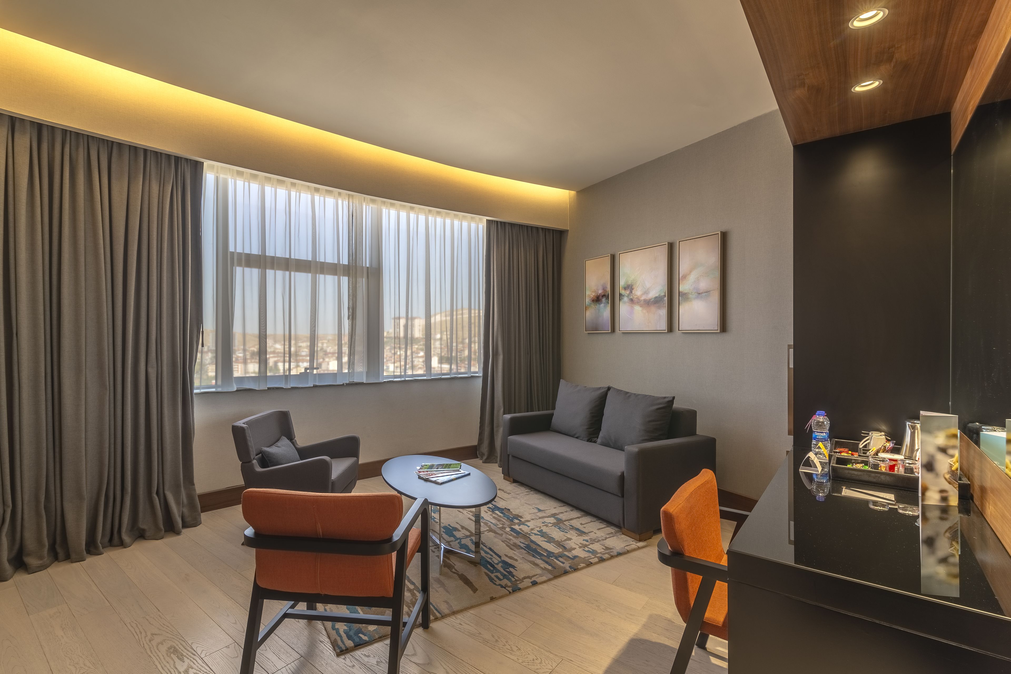 Una habitación de hotel moderna con sofá, dos sillones, una mesita redonda y un amplio ventanal con cortinas.