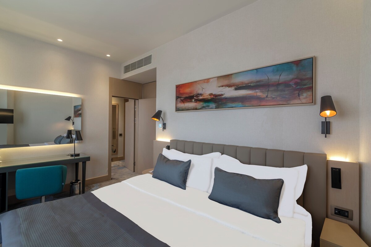 Een moderne hotelkamer met een groot bed, een bureau met stoel, een bureaulamp, een wanddecoratie en twee bedlampen.