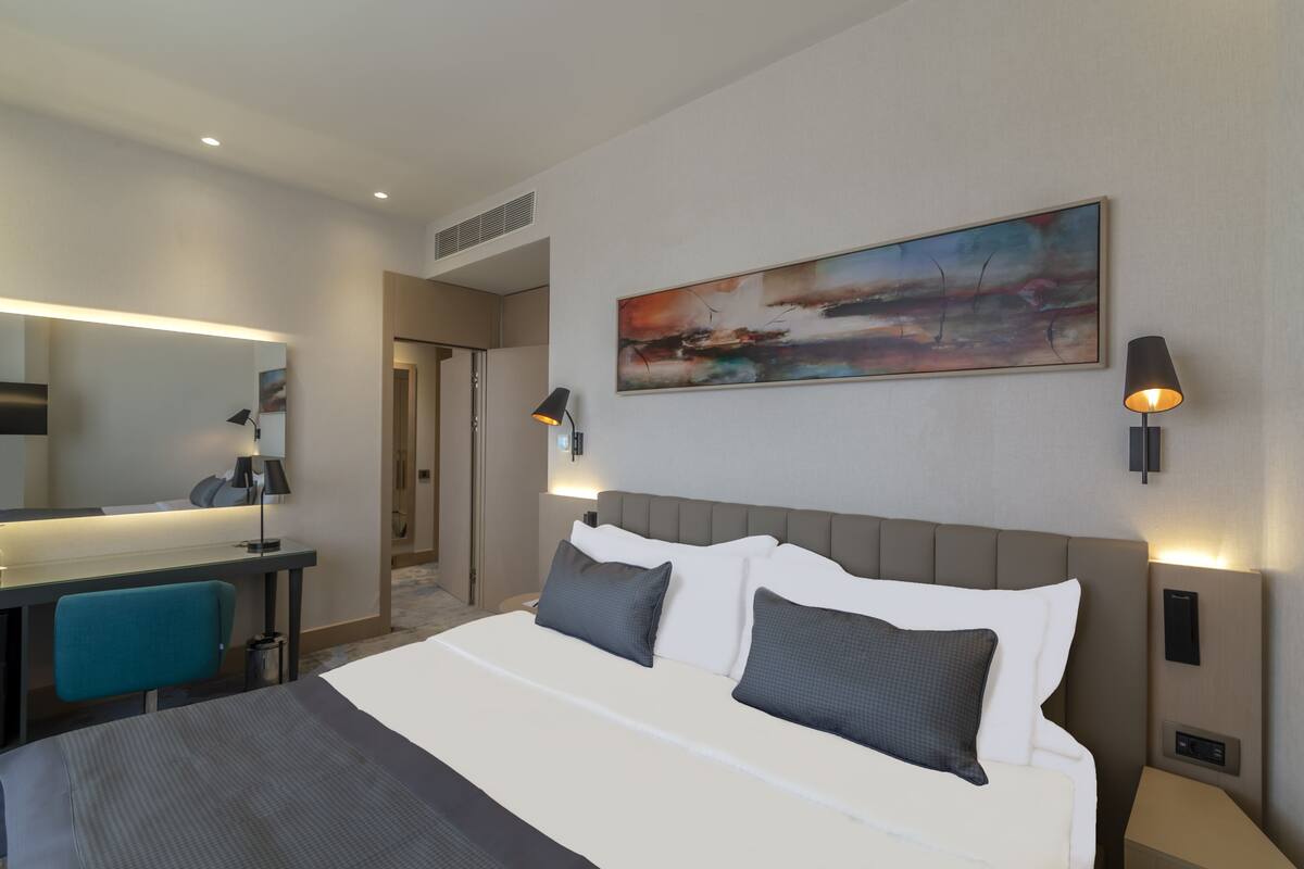 Une chambre d’hôtel avec un grand lit, un bureau et une chaise. Il y a un tableau au mur et deux lampes fixées au mur.