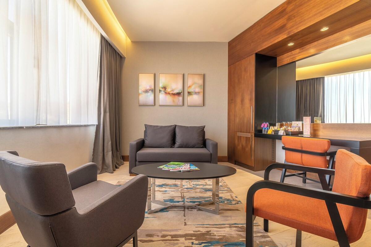 Een moderne hotelkamer met een grijze bank, twee fauteuils, een salontafel en een bureau met een spiegel. Drie abstracte schilderijen hangen aan de muur en er is een raam met gordijnen.