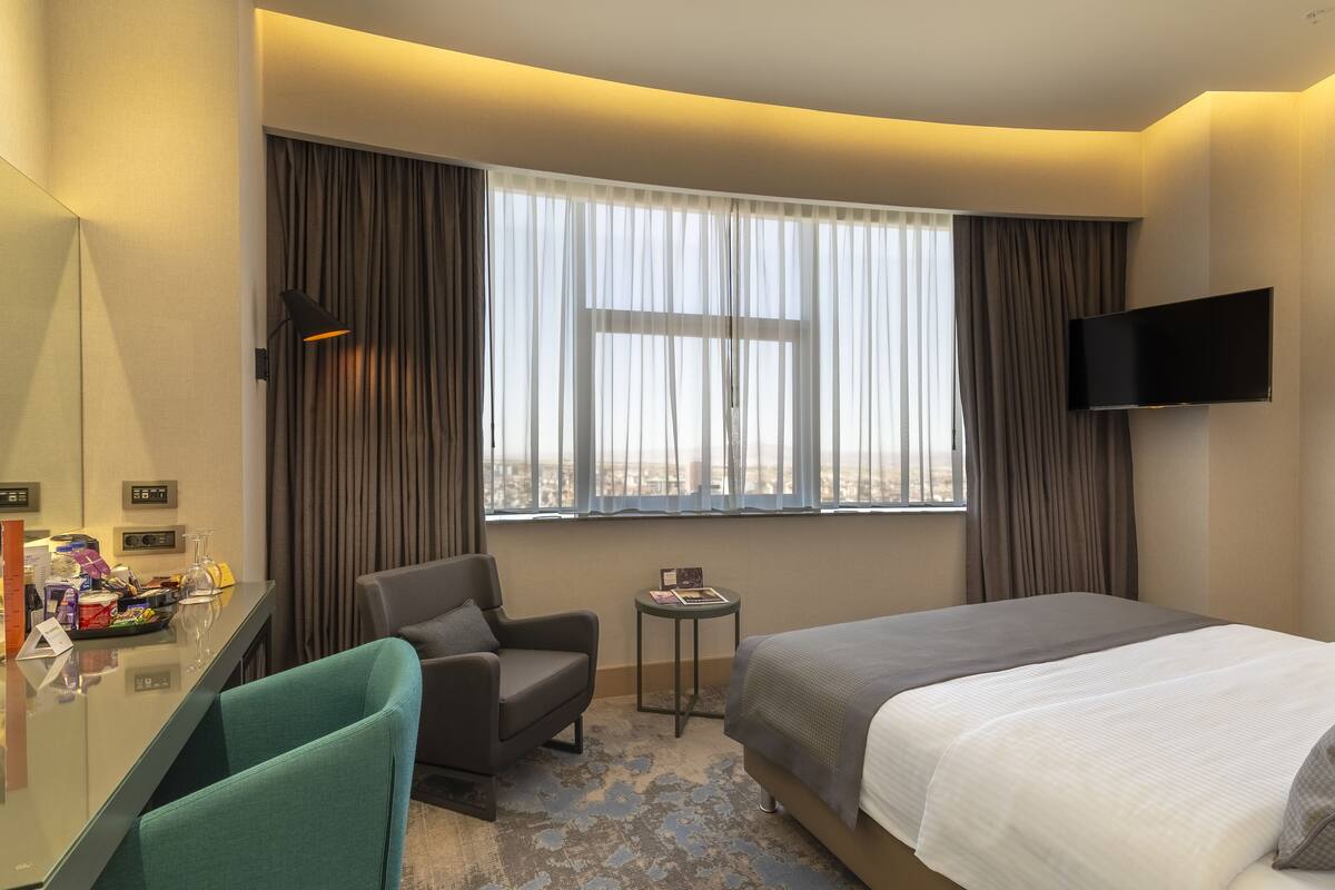 Une chambre d’hôtel avec un lit, un bureau, une chaise, une petite table, un téléviseur et une fenêtre avec des rideaux.