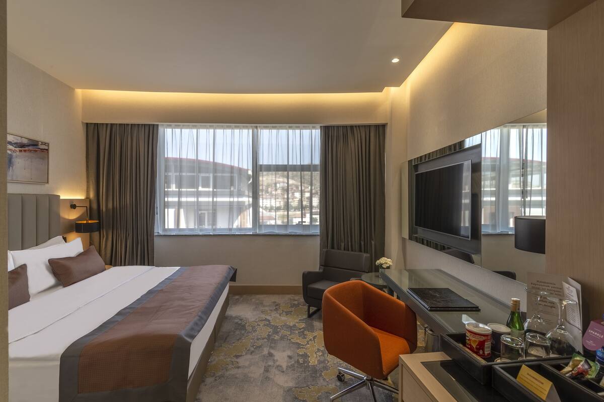 Une chambre d’hôtel avec un lit, un bureau et une chaise, une télévision et une fenêtre avec des rideaux.