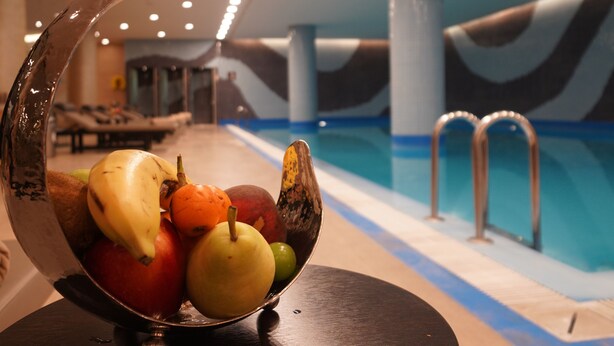 Een schaal met fruit op een tafel, nabij een zwembad met ligbedden en een golvend plafond.