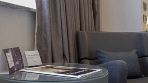 Een moderne hotelkamer met een glazen salontafel, een tijdschrift, een bordje 'Niet roken', een grijze bank en een groot raam met gordijnen.