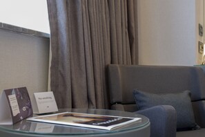 Een moderne hotelkamer met een glazen salontafel, een tijdschrift, een bordje 'Niet roken', een grijze bank en een groot raam met gordijnen.