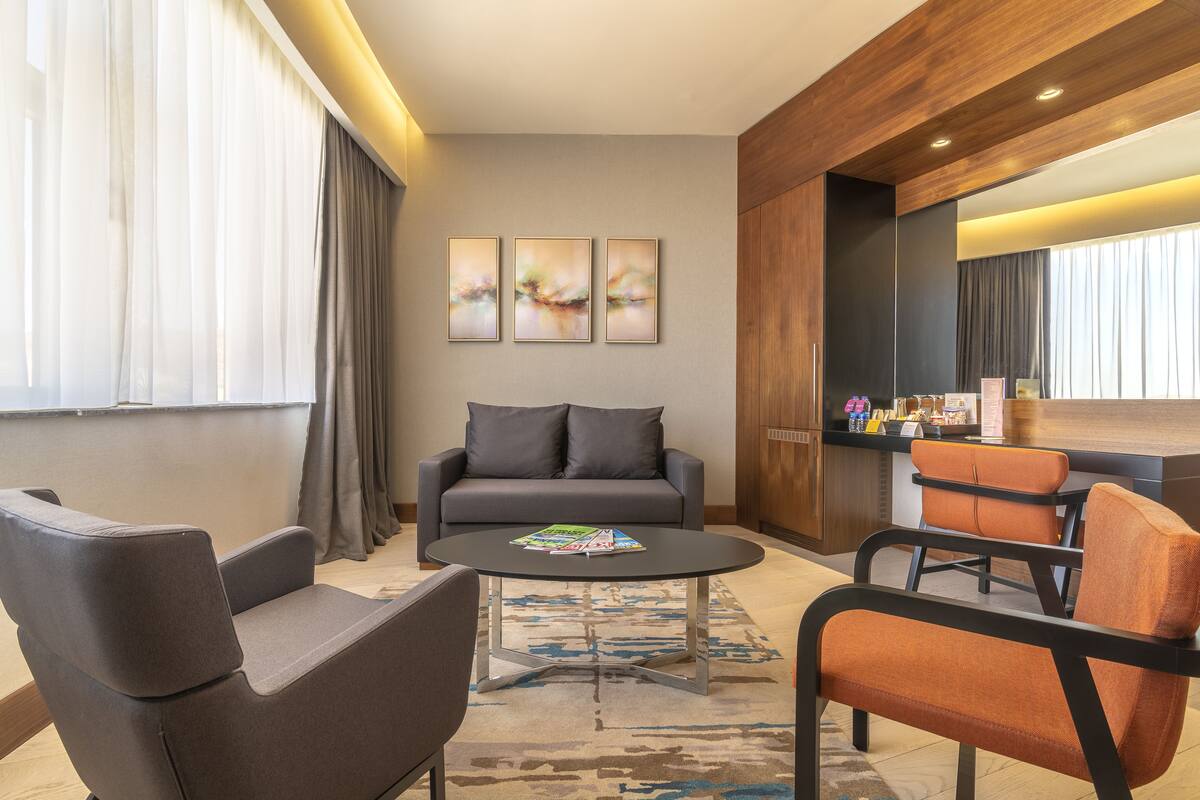 Une chambre d’hôtel moderne avec un canapé gris, deux fauteuils, une table basse et un grand miroir. Trois tableaux abstraits ornent le mur, et un minibar propose divers articles.