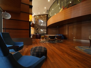 Um interior moderno com uma parede curva de madeira, uma área de jantar com mesa e cadeiras, e uma área de estar com cadeiras azuis e um banquinho xadrez.