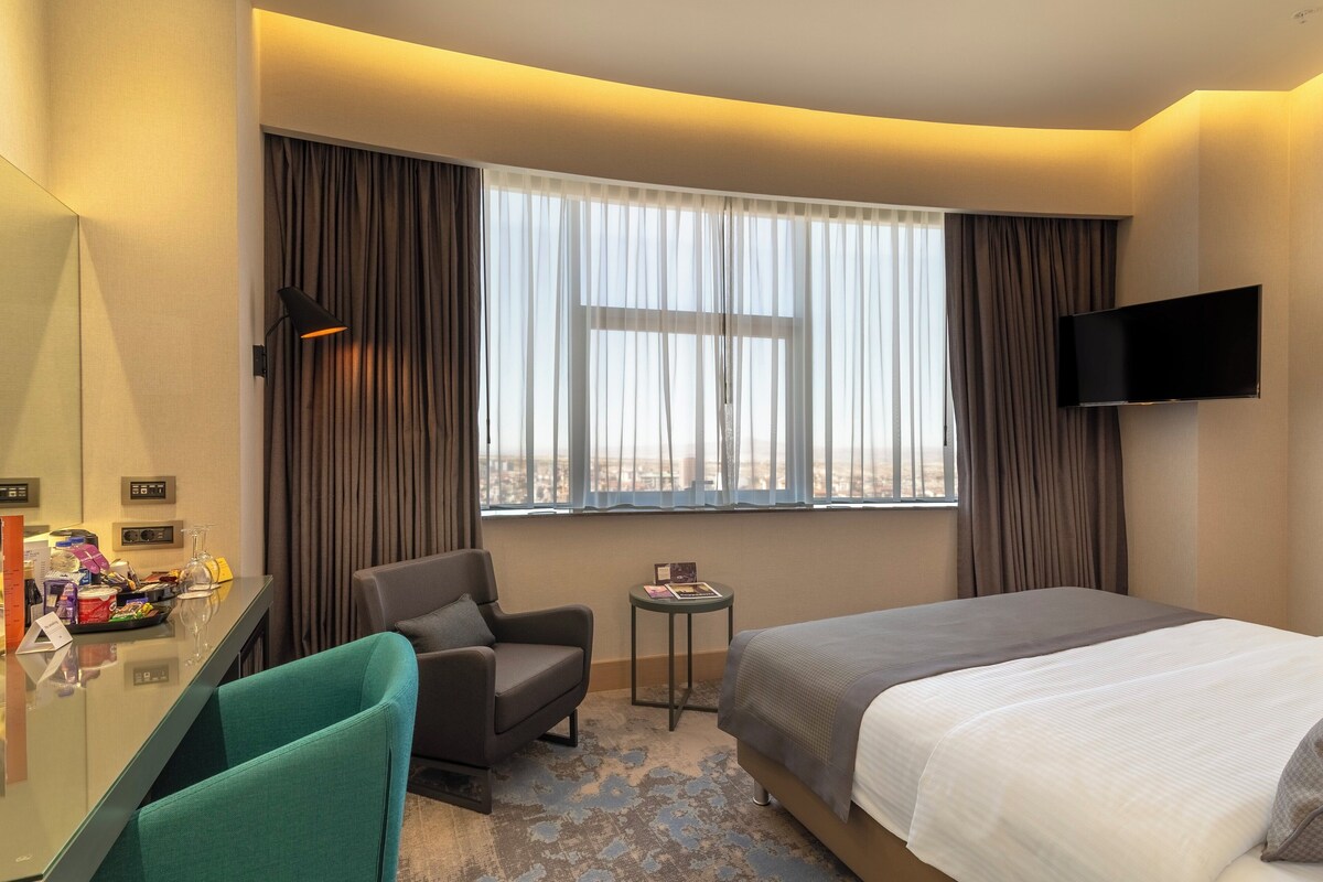 Een hotelkamer met een bed, een bureau, een stoel, een bijzettafeltje, een televisie en een raam met gordijnen.