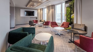 A modern lounge area with green and red upholstered chairs, a flat-screen TV mounted on the wall, and a large window with a view of the city. - Holiday Inn Express Munich - City East by IHG (Munich)