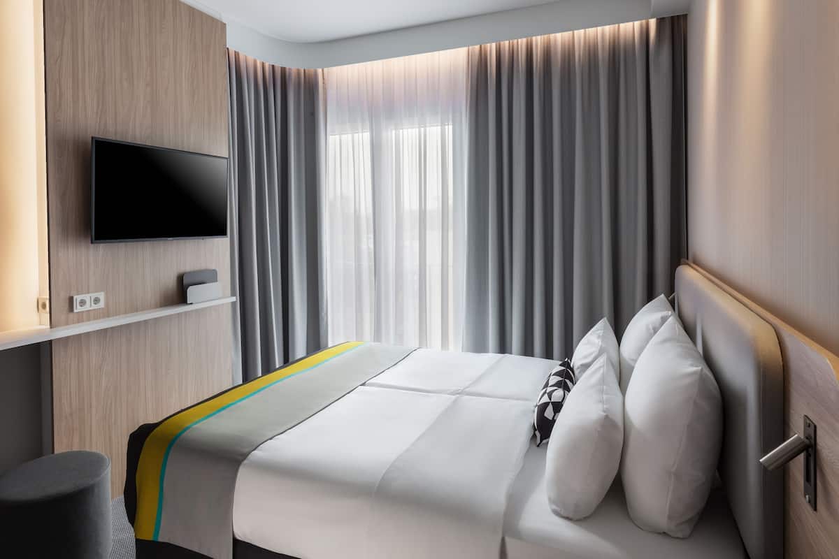 Une chambre d’hôtel moderne dotée d’un téléviseur à écran plat fixé sur un panneau en bois, d’un lit avec du linge de lit blanc et des coussins décoratifs, d  ’une table de chevet avec une lampe, et d’une grande fenêtre ornée de rideaux légers.