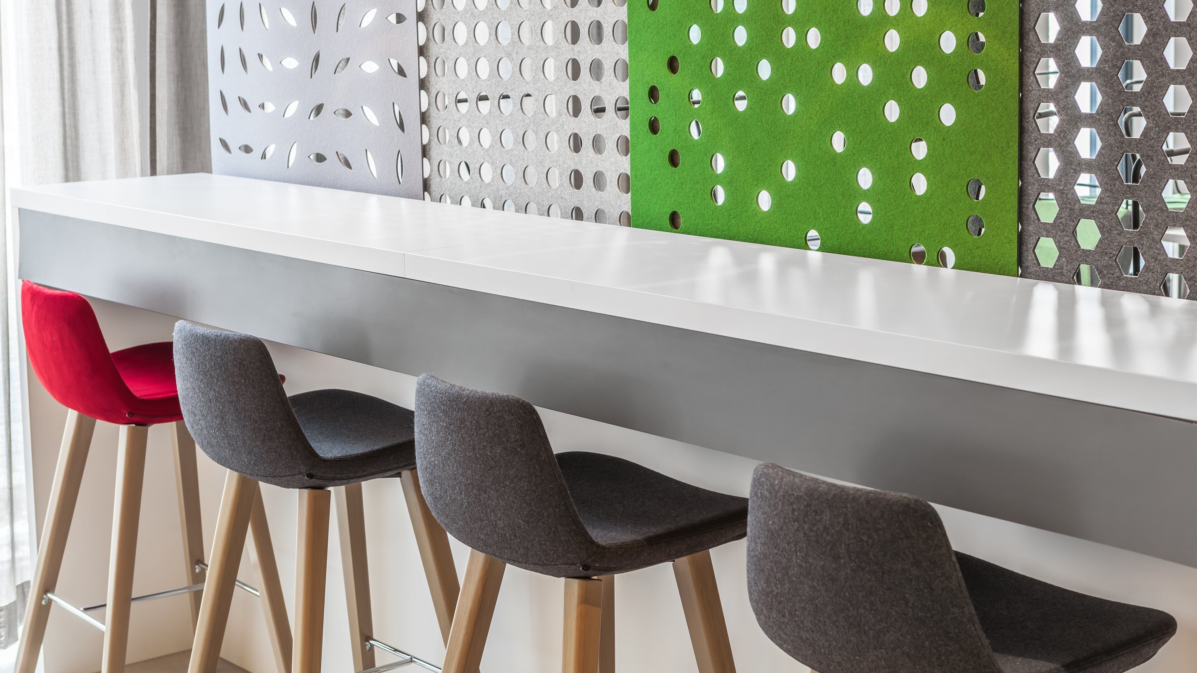 A modern bar area with a white countertop, three chairs with grey and red upholstery, and decorative panels with circular cutouts.