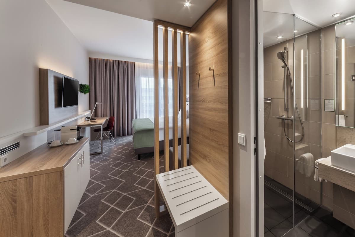Une chambre d’hôtel moderne équipée d’une douche, d’un lavabo, d’un téléviseur fixé au mur et d’un bureau avec une chaise.
