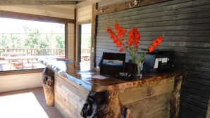 Un mostrador de recepción de madera con diseño rústico, un florero de vidrio con flores rojas, y vistas a la terraza exterior y árboles.