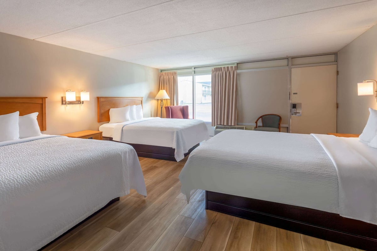 A hotel room with three beds, a wooden floor, a large window with curtains, and a desk with a chair.