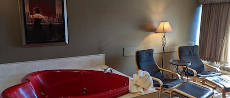 A hotel room with a red jacuzzi tub, a framed painting, a lamp, and two lounge chairs.