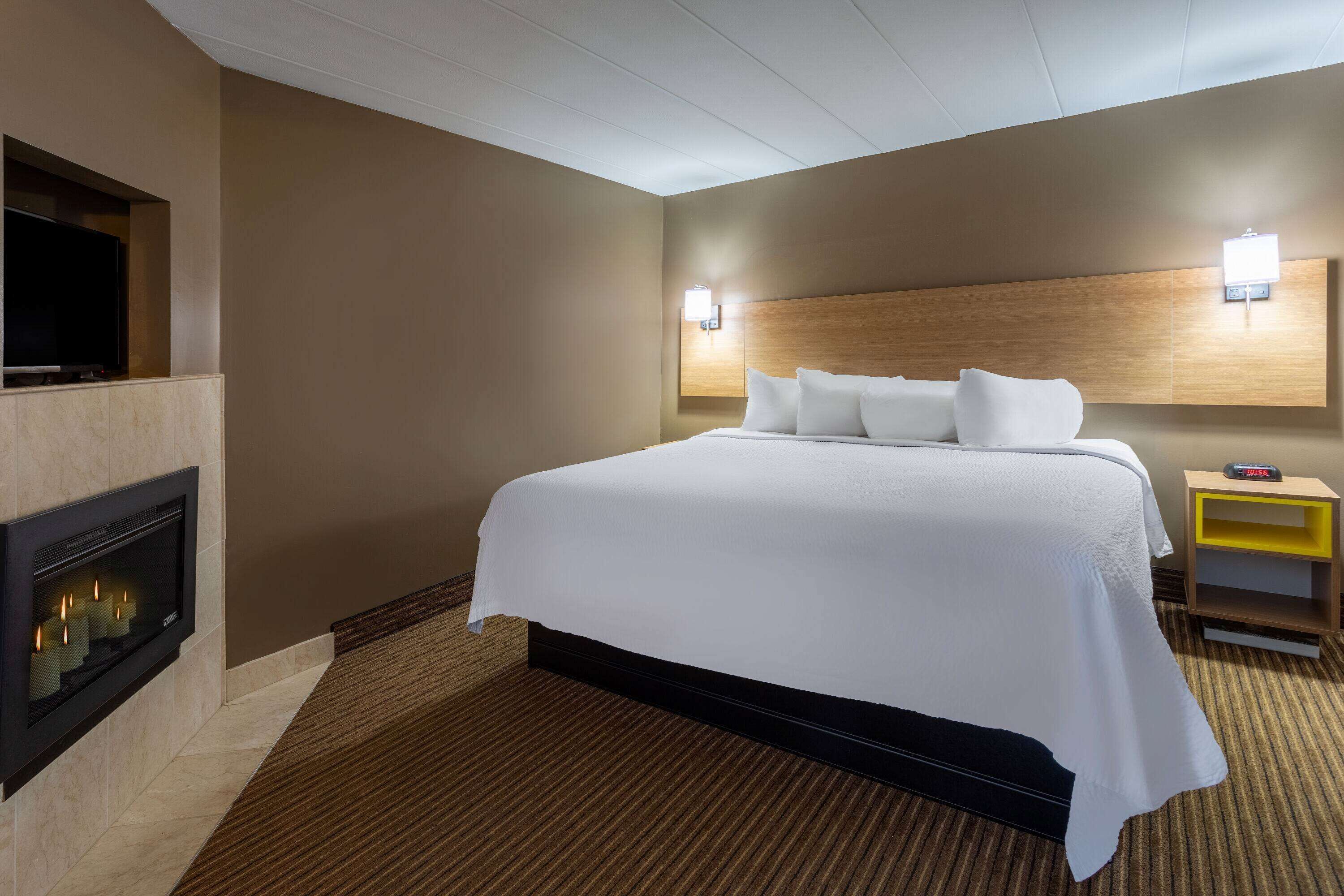 Habitación de hotel con una cama grande, televisión montada en la pared, chimenea y mesita de noche con un reloj.
