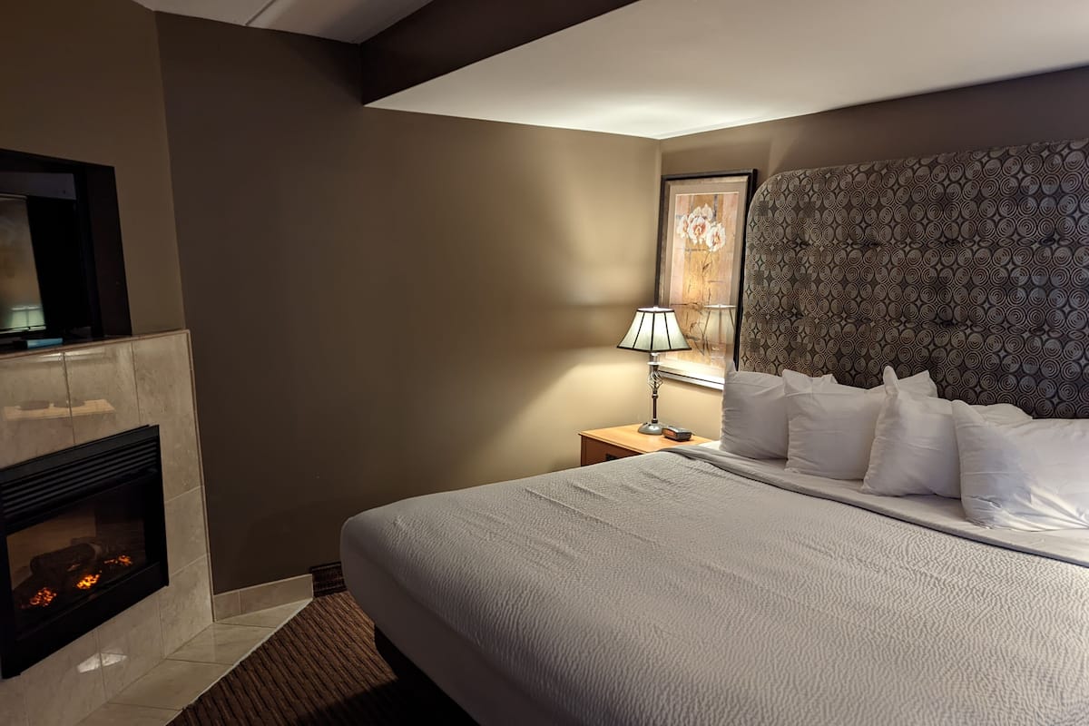 A hotel room with a large bed, a fireplace, a nightstand with a lamp, and a wall-mounted TV.