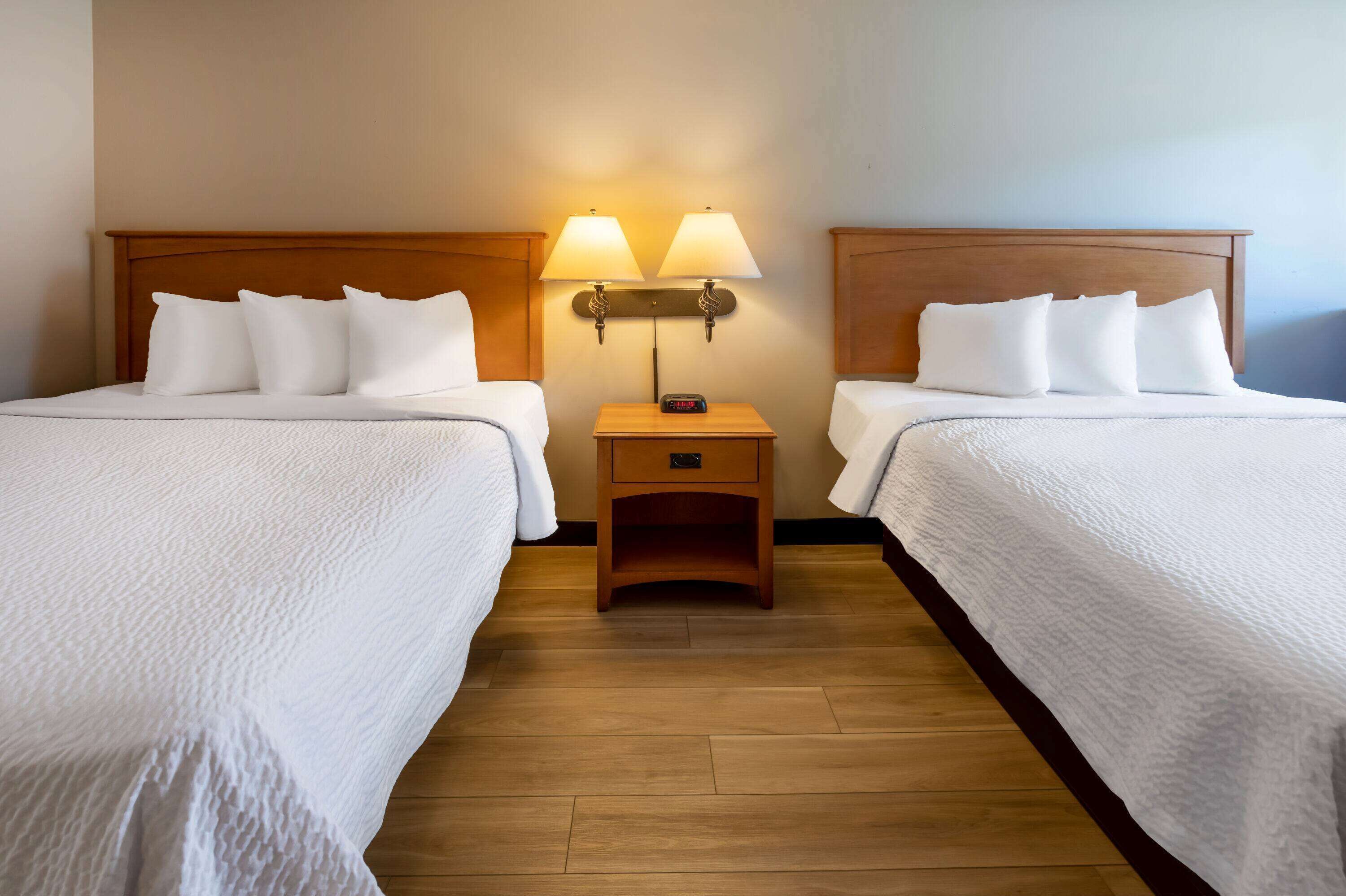 A hotel room with two beds, each with white linens and pillows, a wooden headboard, a nightstand with a lamp, and a small drawer.