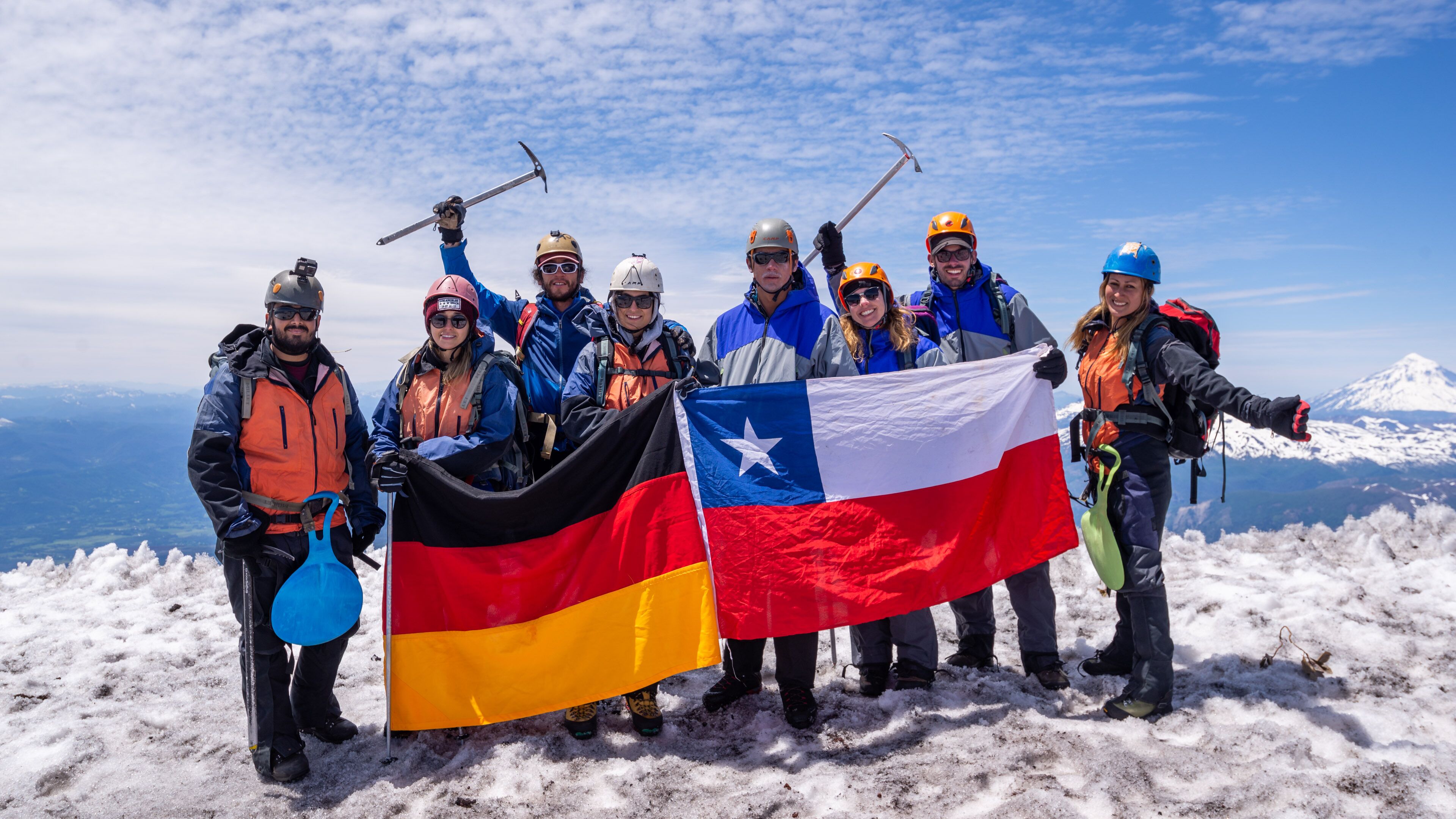Un grupo de personas en una montaña nevada con banderas, usando cascos y cargando mochilas.