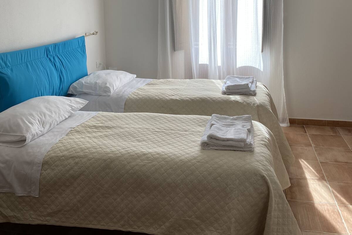 Une chambre d’hôtel avec deux lits, chacun recouvert d’une courtepointe matelassée et agrémenté d’oreillers blancs. Une grande fenêtre est ornée de rideaux légers, et un panneau de tête de lit en bois se trouve au-dessus des lits.
