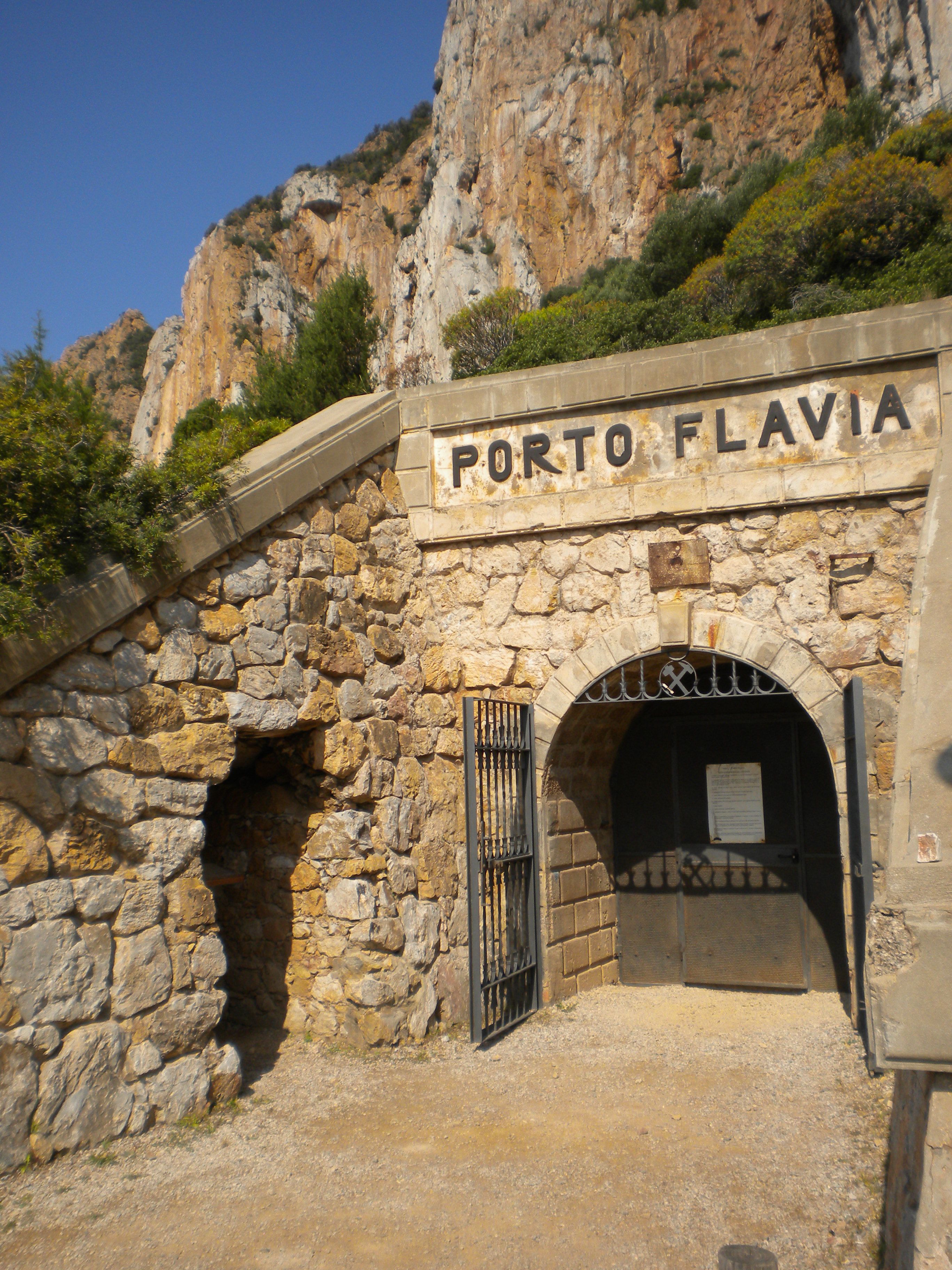 A stone building with a sign reading "PORTO FLAVIA" and a metal gate.