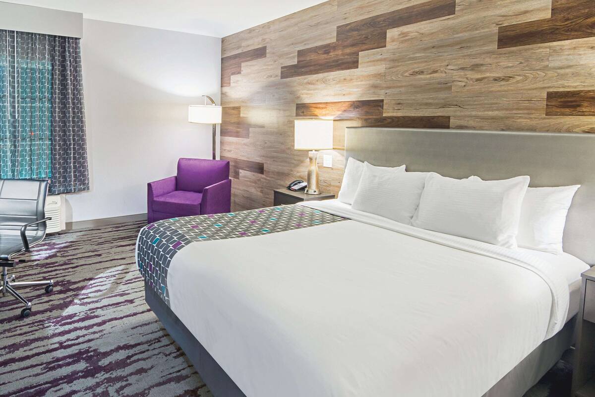 Een hotelkamer met een groot bed, een paarse fauteuil, een bureau met een lamp en een kamerbreed tapijt met dessin.