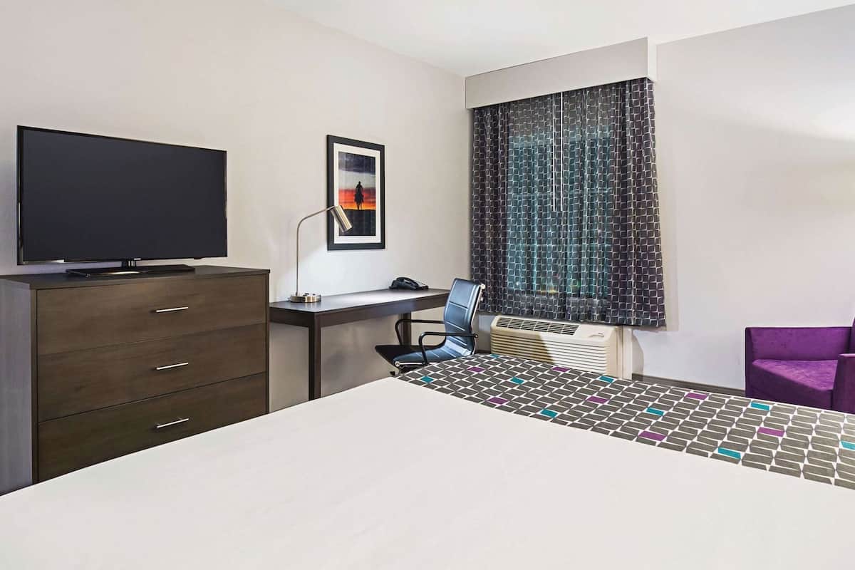 Een hotelkamer met een bed, een bureau met stoel, een televisie, een ingelijst beeld en een raam met gordijnen met dessin.