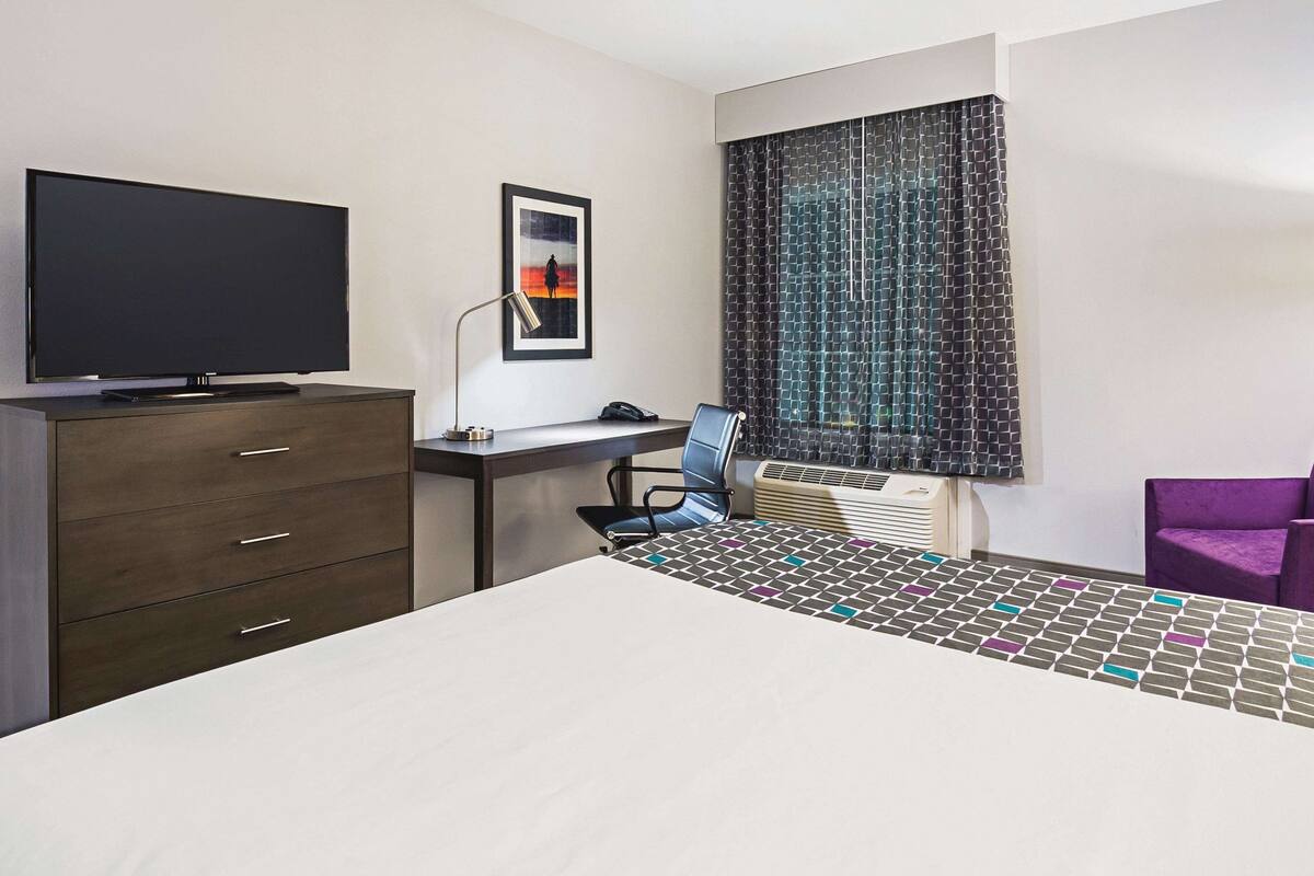 Hotelkamer met een bed, een bureau met een computer, een stoel, een televisie en een raam met gordijnen met een patroon.