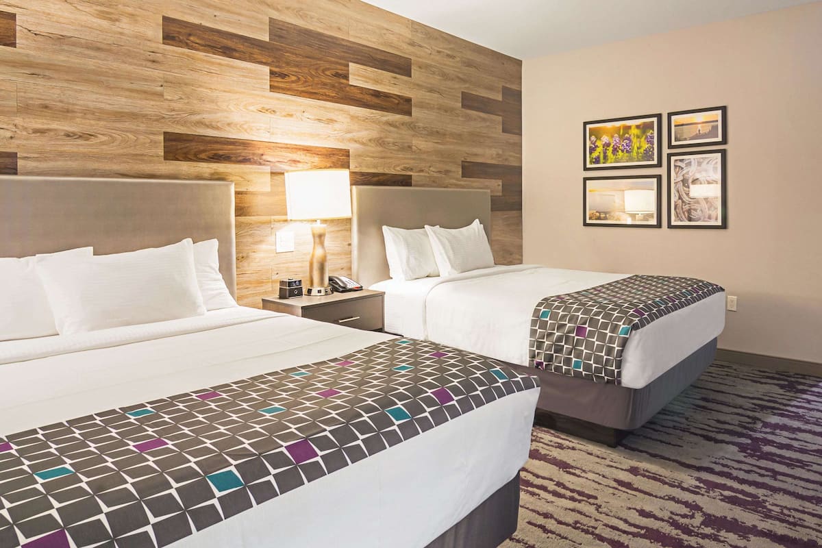 Hotelkamer met twee bedden, houten hoofdeinden, een bed met een decoratieve spreidek en een nachtkastje met een lamp, plus kunstwerken in een lijst aan de muur.