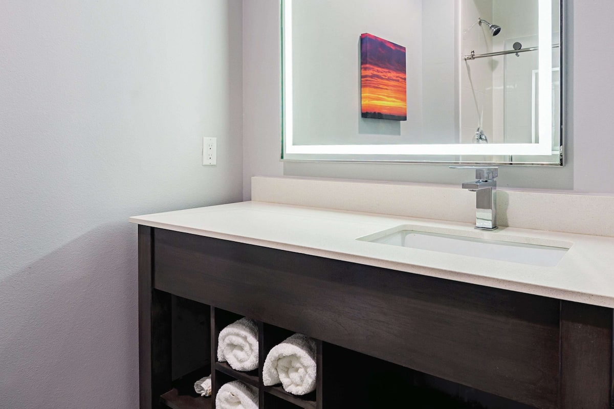 Een moderne badkamer met een witte wasbak, een donker houten opbergmeubel en een glazen plank met opgerolde handdoeken.
