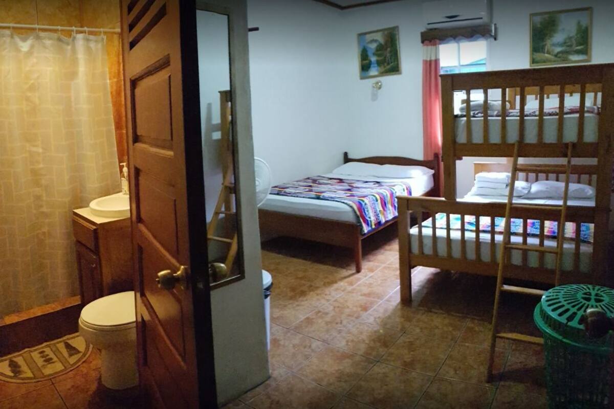 A room with a wooden bed, a small bedside table, a window with curtains, a bathroom with a toilet and sink, and a green trash bin.