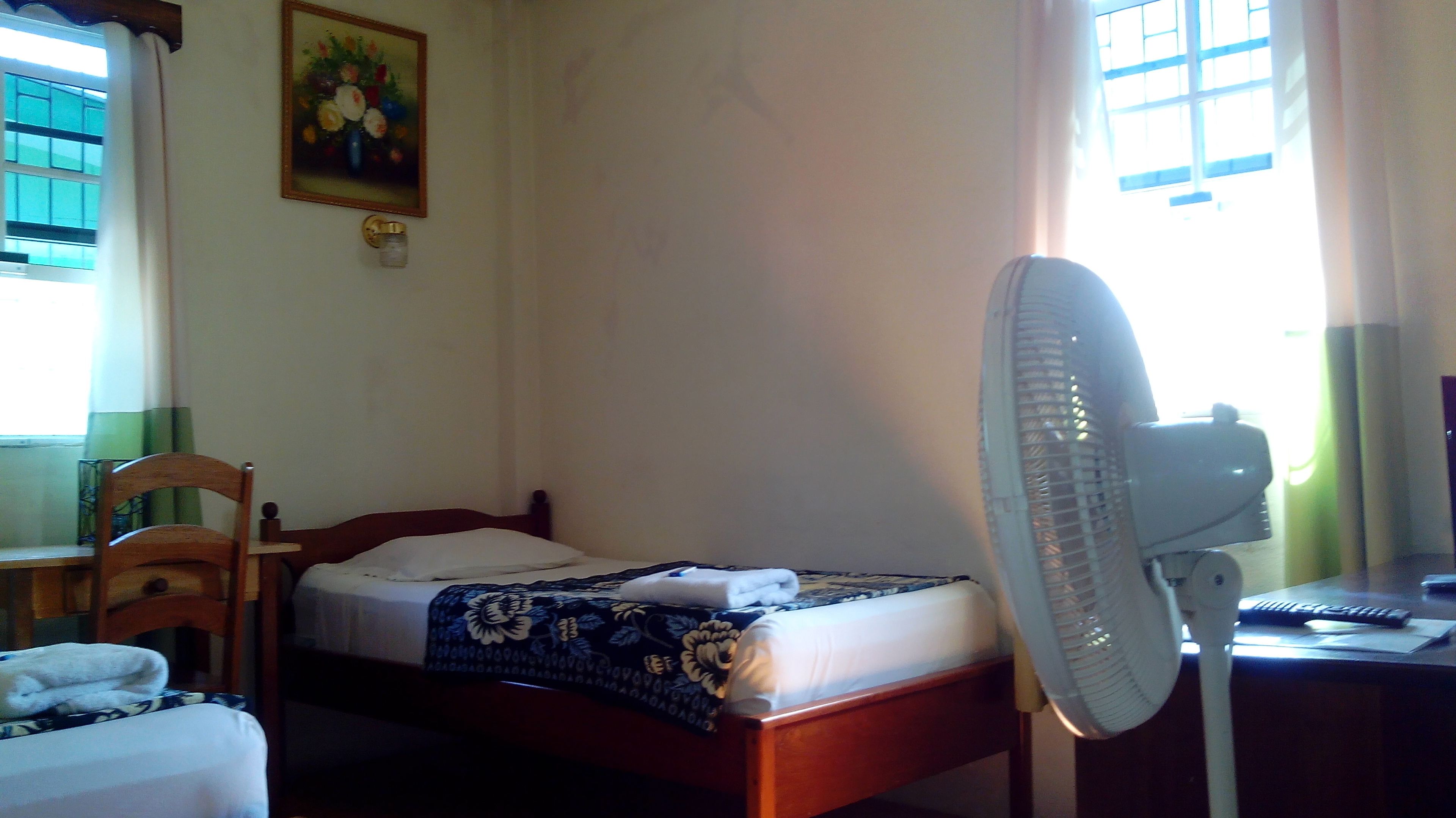 A bedroom with a bed, a desk, a chair, a standing fan, and a window with curtains.