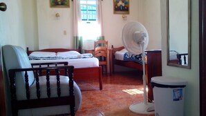 Quarto com cama, uma cadeira, um ventilador e uma lixeira.