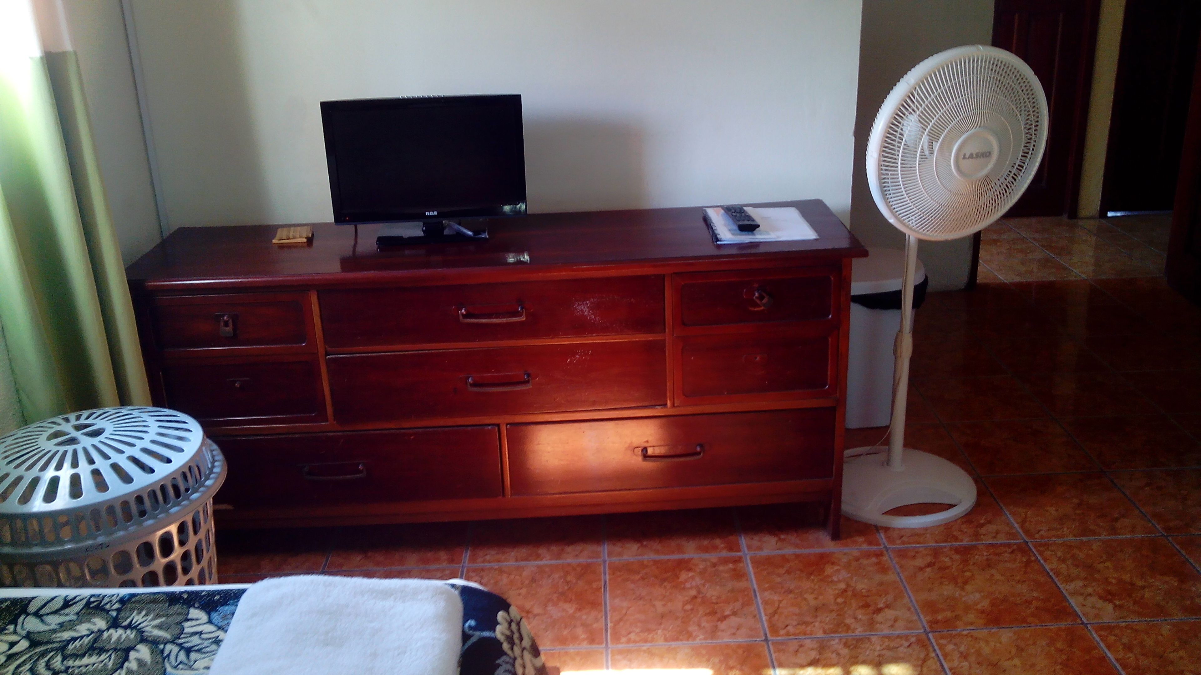 Une commode en bois, un téléviseur à écran plat, une télécommande et un ventilateur sur pied blanc.