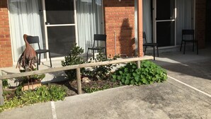 A building with a brick facade, large windows with curtains, and a concrete patio area with chairs and plants. - Inverloch Central Motor Inn (Inverloch)