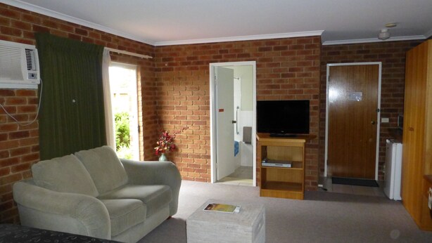 A living room with a sofa, a television on a wooden stand, a brick wall, and a door leading to another room. - Inverloch Central Motor Inn (Inverloch)