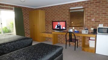 A room with a brick wall, a wooden desk with a computer, a chair, a bed, and a small kitchenette.