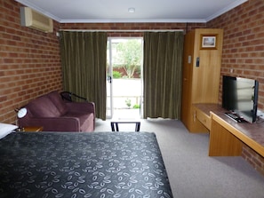 Habitación de hotel con una cama, un escritorio con televisor, una silla y un ventanal con cortinas.